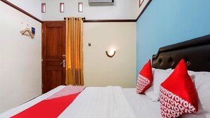 A hotel room with a bed, a wooden door, a curtain, a wall-mounted light, and a red pillow with a pattern. - OYO 3892 Athaya Guesthouse Syariah (Depok)