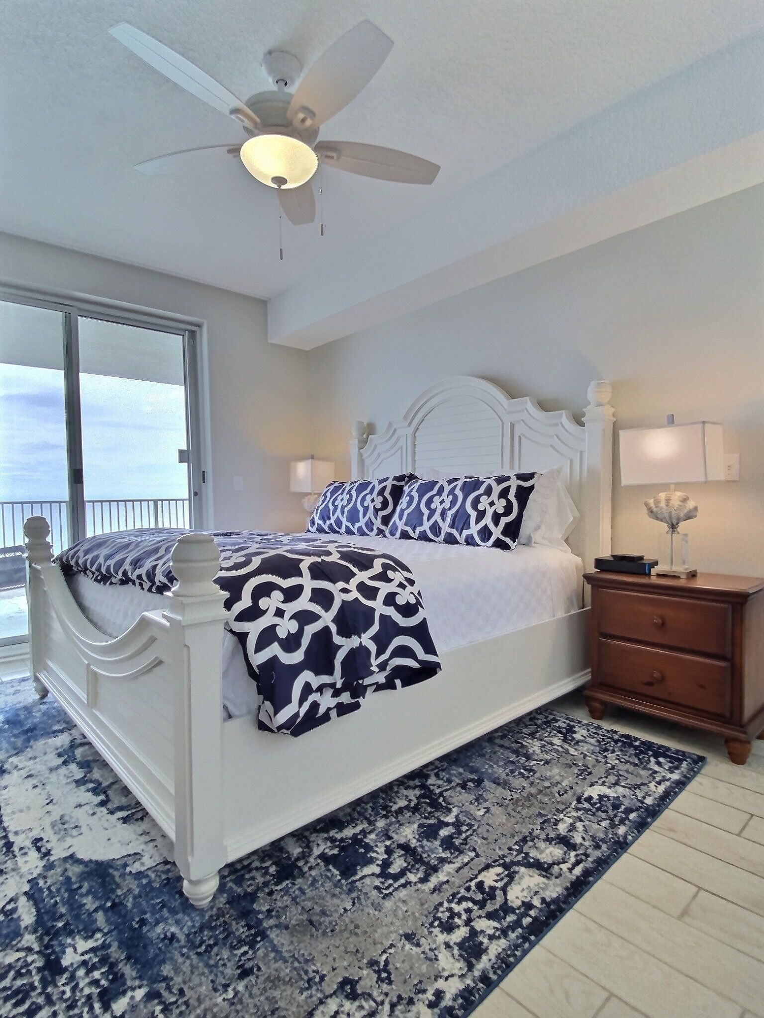 A bedroom with a white bed, patterned bedding, a ceiling fan, a nightstand, and a view through a sliding glass door.