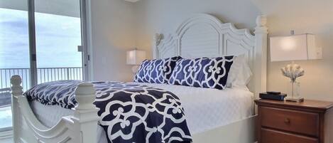 A bedroom with a white bed, patterned bedding, a ceiling fan, a nightstand, and a view through a sliding glass door.