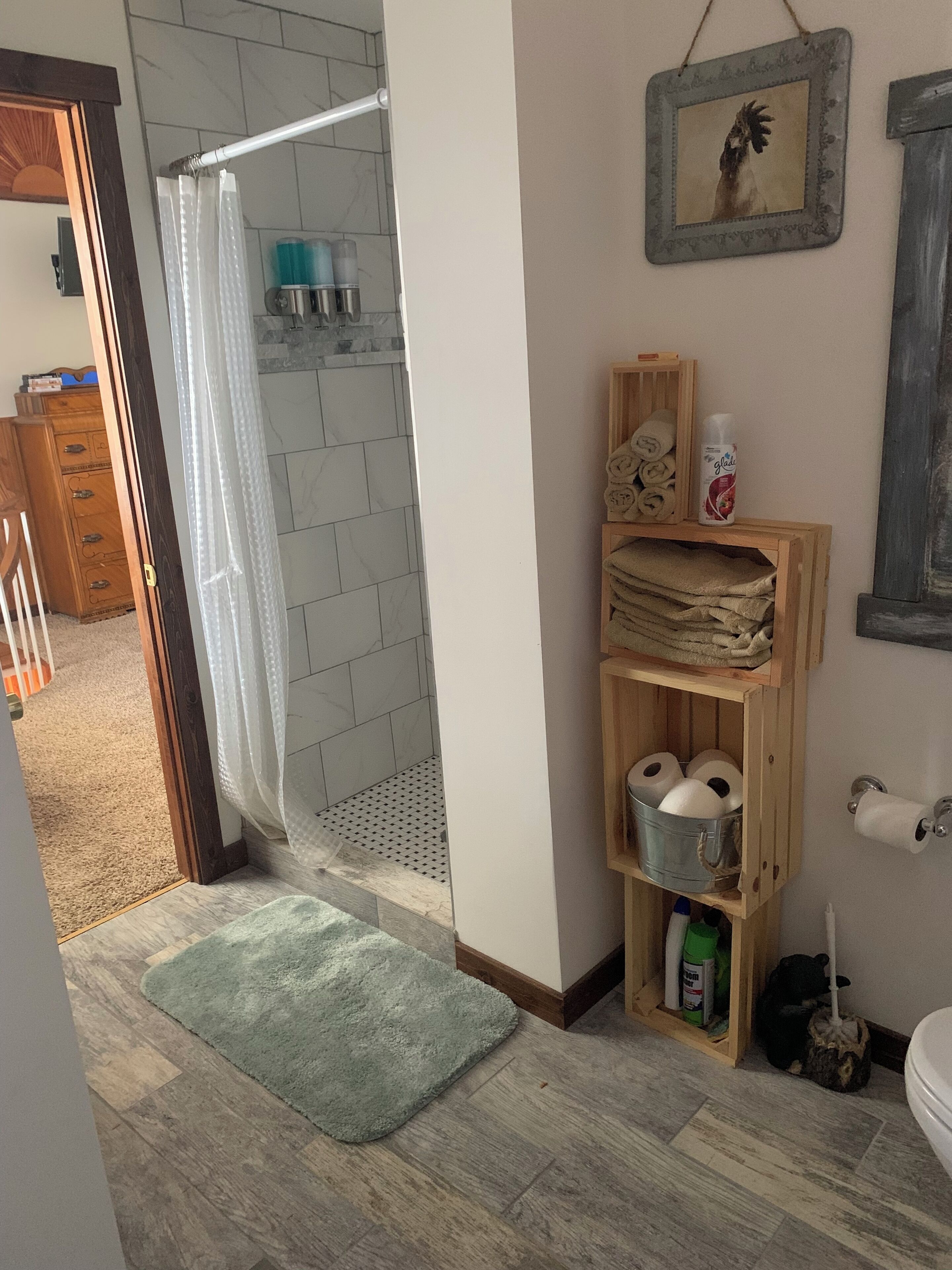 A bathroom with a shower, toilet paper, and a wooden shelf with towels and toiletries.