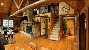 Un intérieur spacieux de chalet en bois, avec une cheminée, un canapé, une table à manger et un escalier menant à un niveau supérieur.