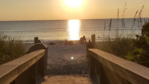 Une promenade en bois qui mène à une plage, avec le soleil couchant en arrière-plan.