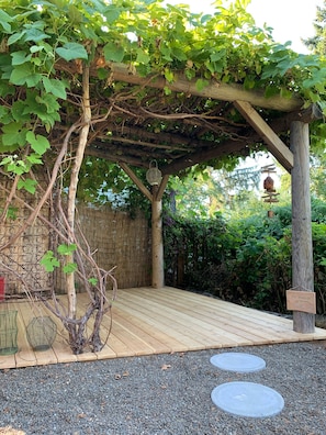 En træterrasse med en pergola dækket af slyngplanter, omgivet af et bambushegn og frodig vegetation.