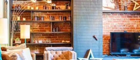 A cozy living room with a brick wall, a fireplace, a television, and a bookshelf filled with books and decorative items.
