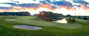 Un parcours de golf avec des bunkers, un obstacle d’eau et un coucher de soleil en arrière-plan.