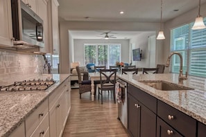 A modern kitchen with granite countertops, a gas stove, and dark cabinetry. There is a dining area with a table and chairs, and a ceiling fan. - Bayside * 5BR Luxury Twin House (Selbyville)
