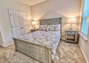 Une chambre à coucher comprenant un lit, deux tables de chevet, une commode, une porte et une fenêtre avec des stores.