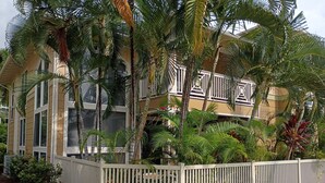 Un immeuble à plusieurs étages doté de balcons, entouré d’une végétation tropicale luxuriante et d’une clôture blanche.