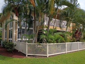 Un edificio a più piani con balconi, immerso nel verde di piante tropicali e circondato da un recinto bianco.