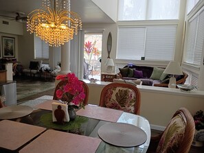 Zona pranzo con tavolo in vetro, sedie, un lampadario e vista sull'esterno attraverso ampie finestre.