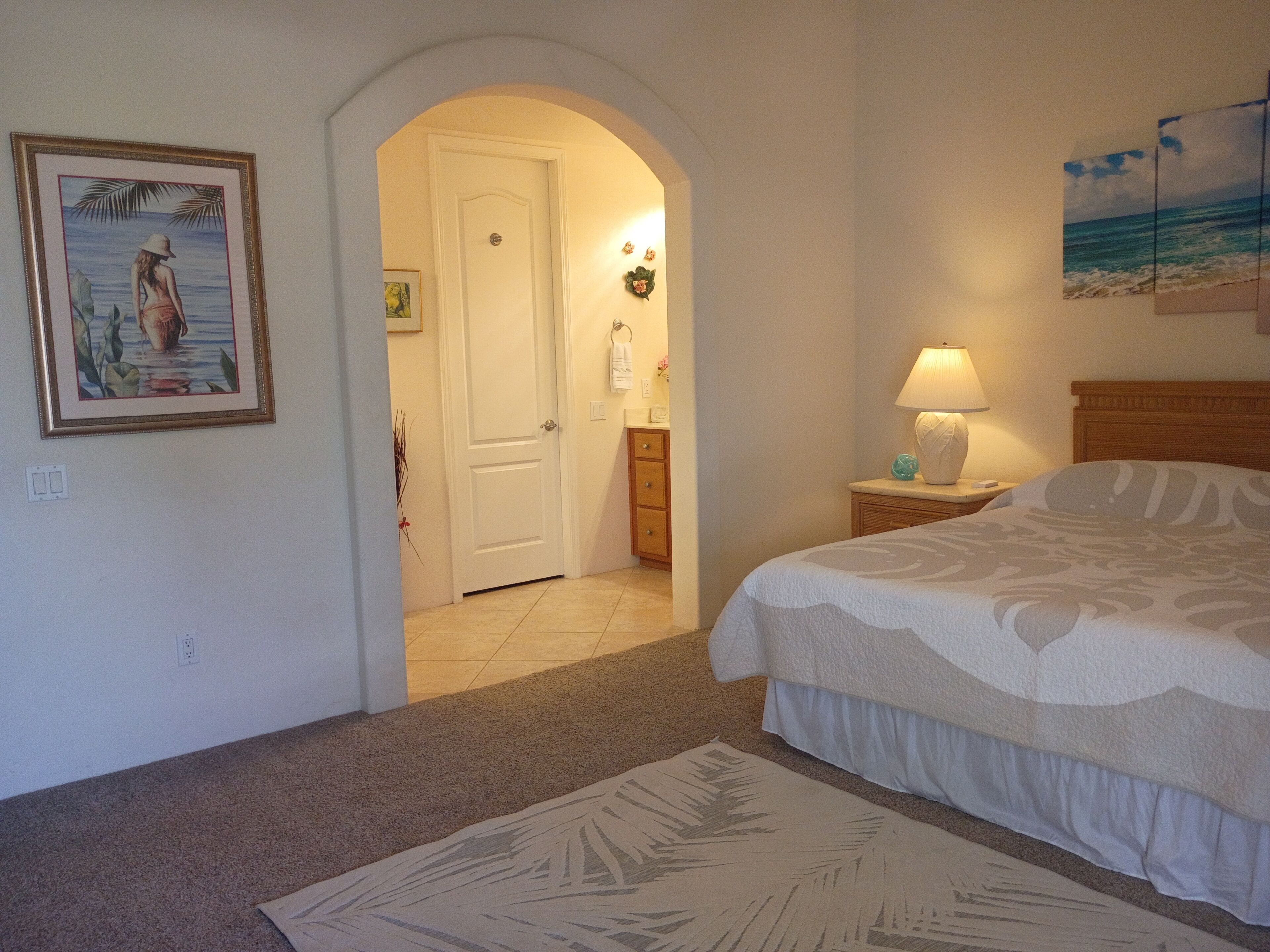 Un dormitorio con cama, mesita de noche, lámpara, un cuadro enmarco y un arco que da acceso a un baño.