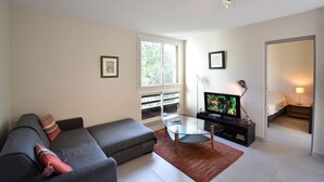 A modern living room with a sofa, a glass coffee table, a television, and a balcony with a view of greenery. - Seignosse “LE PIGNADA” Apartment for 4 people. (SEIGNOSSE)