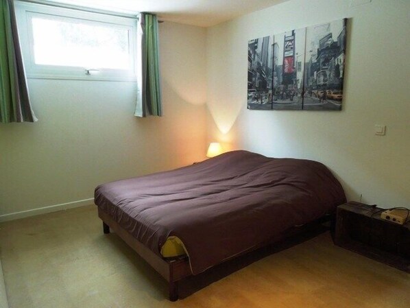 A bedroom with a bed, a window with curtains, a wall-mounted lamp, and a painting.