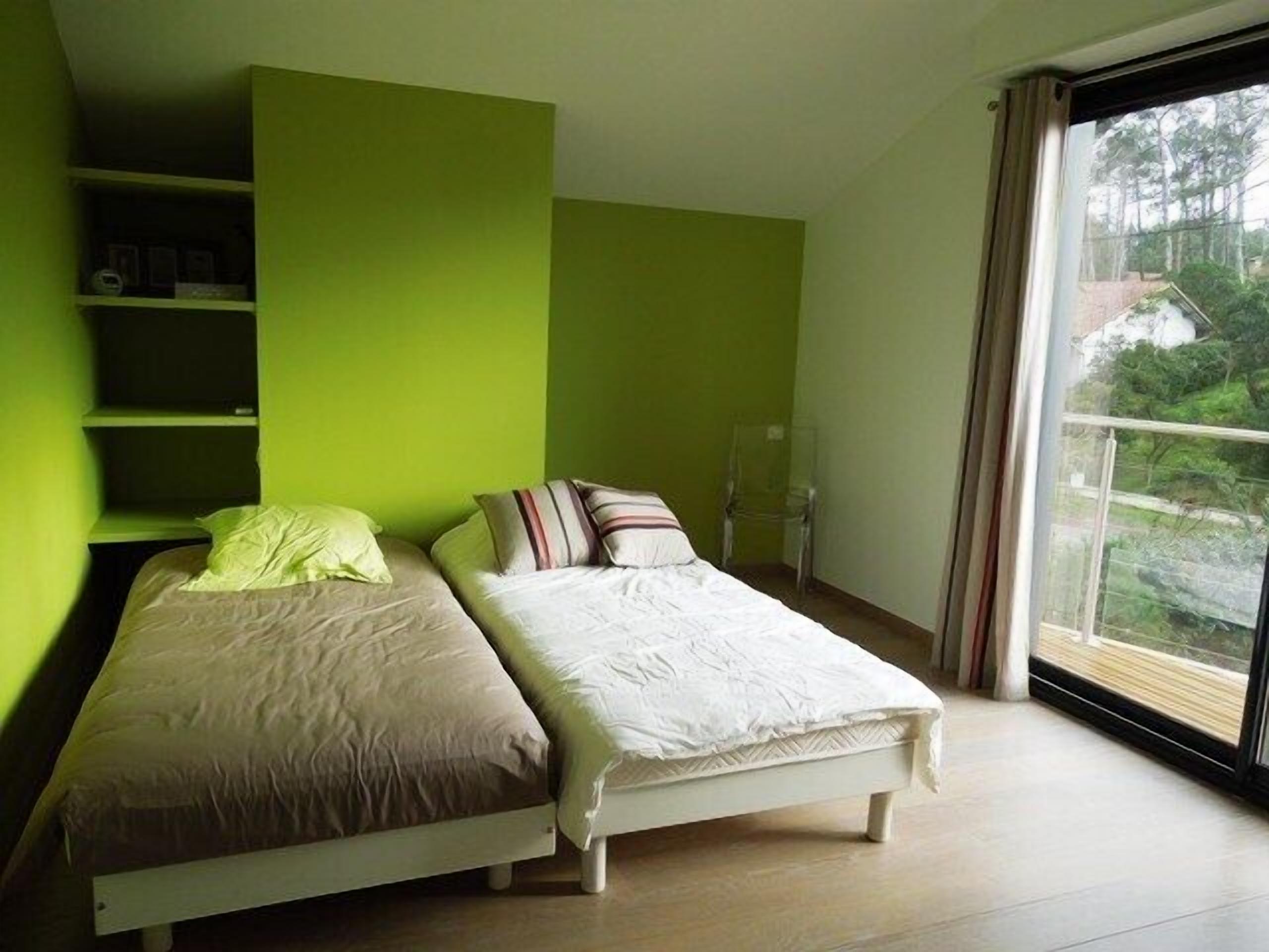 A bedroom with two beds, a green wall, and a large window with a view of trees.
