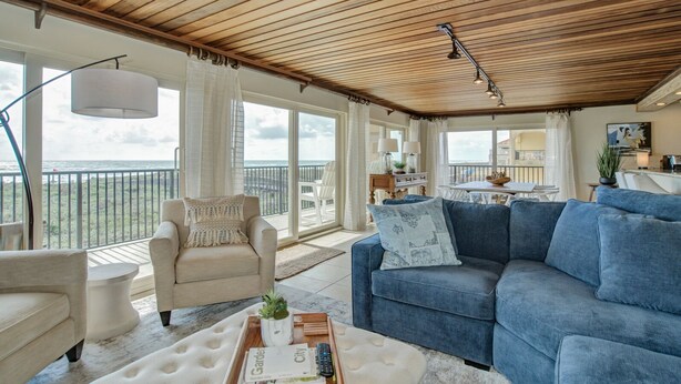 Een ruime woonkamer met een houten plafond, een blauwe bank, een beige fauteuil en een groot raam met uitzicht op de oceaan.