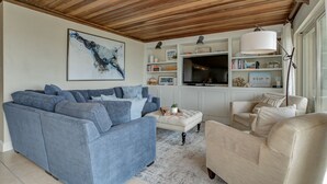 Een woonkamer met een blauwe modulaire bank, een beige fauteuil en een wit voetenbankje. Er is een grote flatscreen televisie op een witte kast, en een houten plafond.