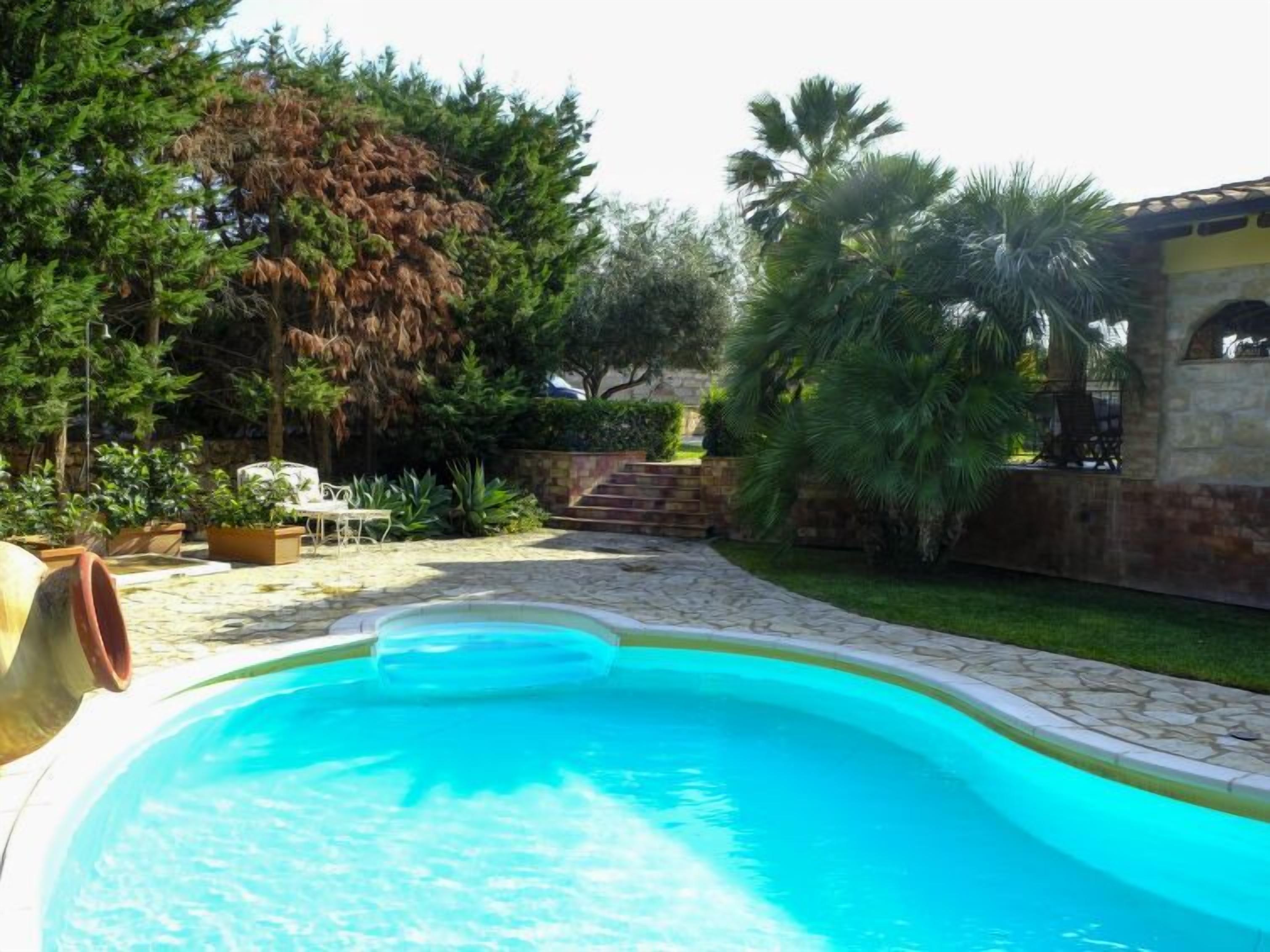 A swimming pool with a stone deck, surrounded by lush greenery and a stone wall.
