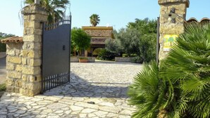 A stone entrance gate leading to a building with a tiled roof, surrounded by greenery and palm trees.