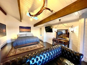 A bedroom with a large bed, a flat-screen TV, a wooden ceiling, and a black leather sofa.