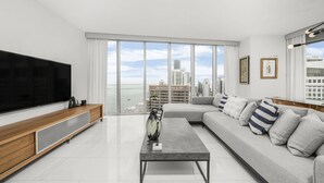 A modern living room with a large TV, a grey sofa, and a view of the city skyline through floor-to-ceiling windows. - ICON W BRICKELL MARVELOUS OCEAN VIEW 2BR/2BA #4508 (Miami)