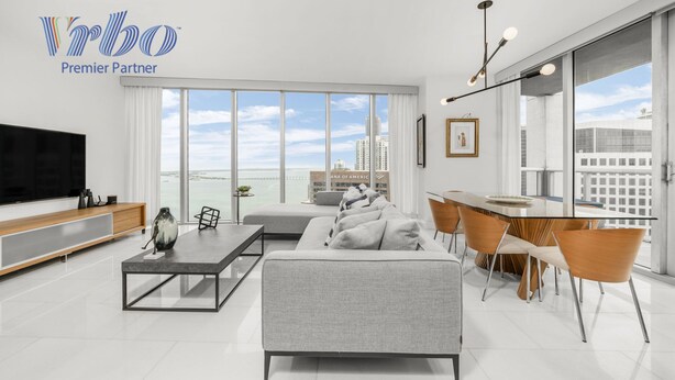 A modern living room with a large TV, grey sofa, dining table with chairs, and large windows offering a city view. - ICON W BRICKELL MARVELOUS OCEAN VIEW 2BR/2BA #4508 (Miami)