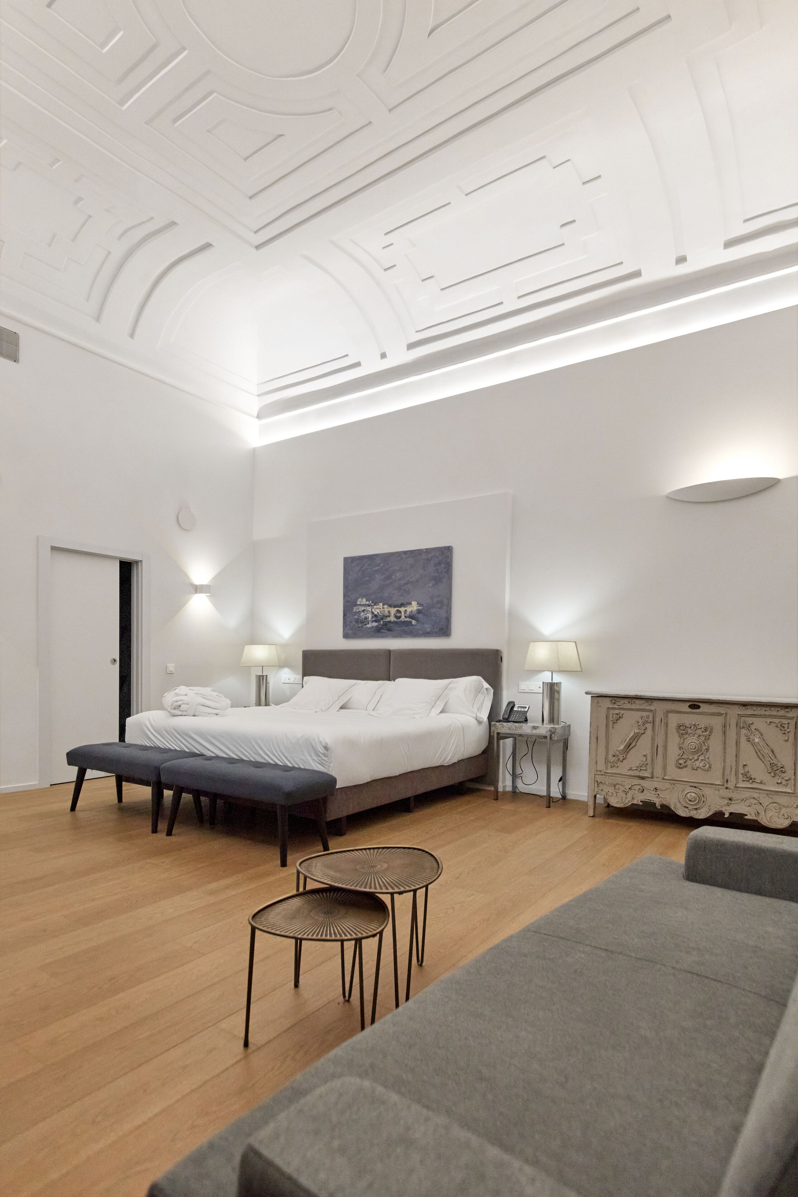 A modern hotel room with a large bed, two bedside tables, a grey sofa, a small round table, and a decorative cabinet.