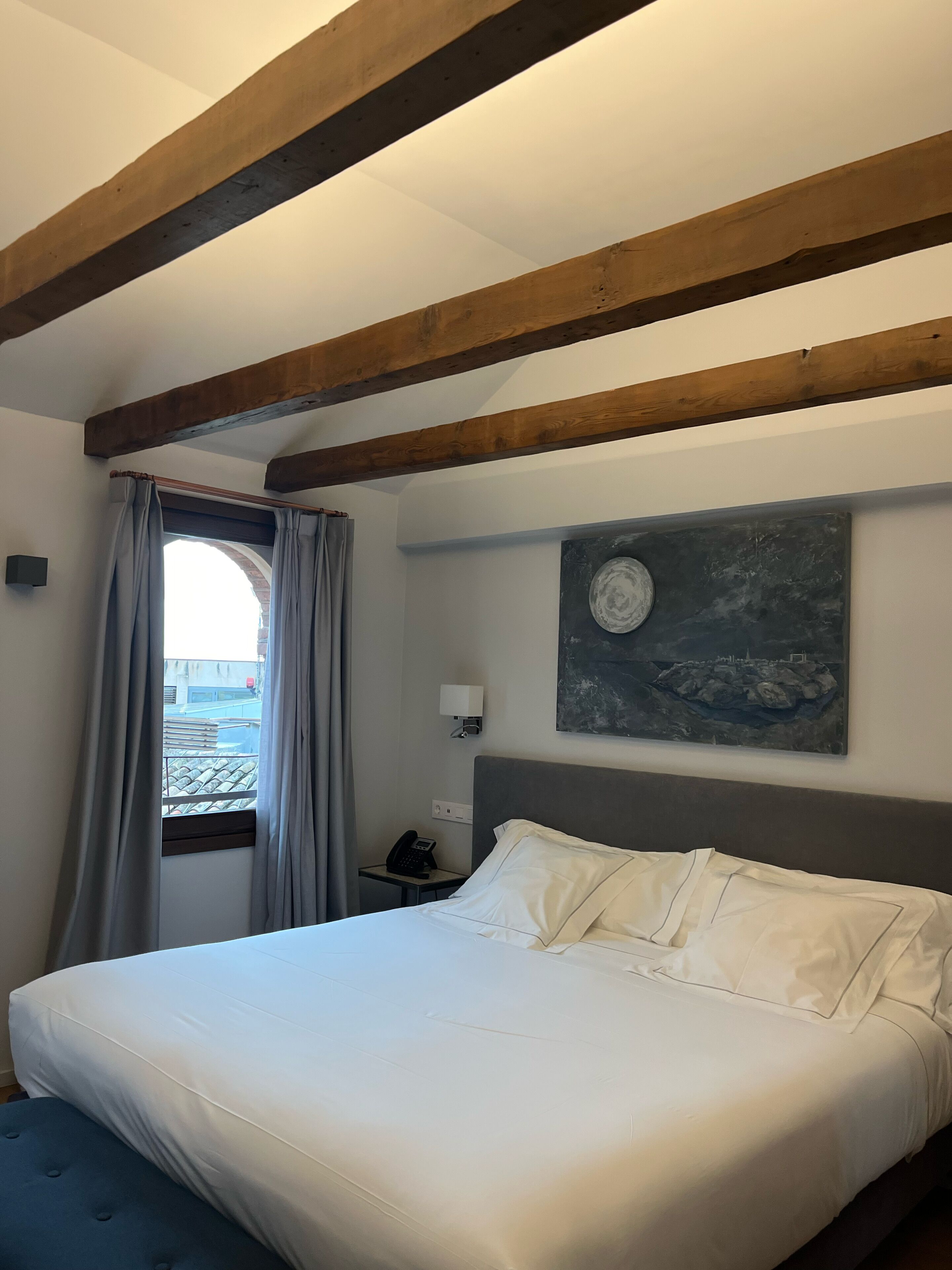 A hotel room with a bed, a nightstand, a painting, and a window with curtains.