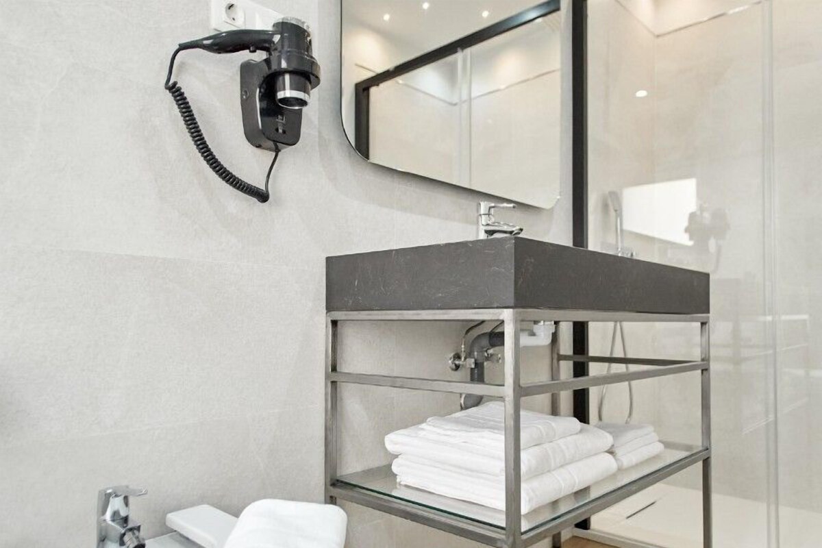 Een moderne badkamer met een glazen plank met opgevouwen handdoeken, een zwarte haardroger en een spiegel boven de wastafel.