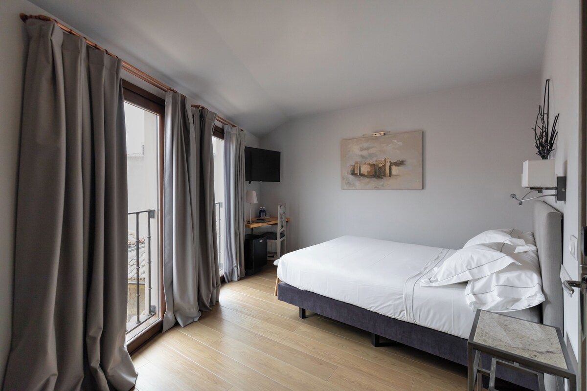 Een slaapkamer met een bed, een bureau, een stoel, een televisie en een raam met gordijnen.