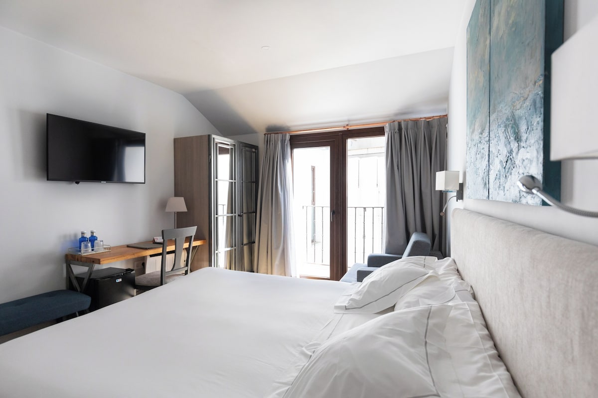 Habitación de hotel con una cama grande, un escritorio con silla, televisión montada en la pared y un balcón con cortinas.