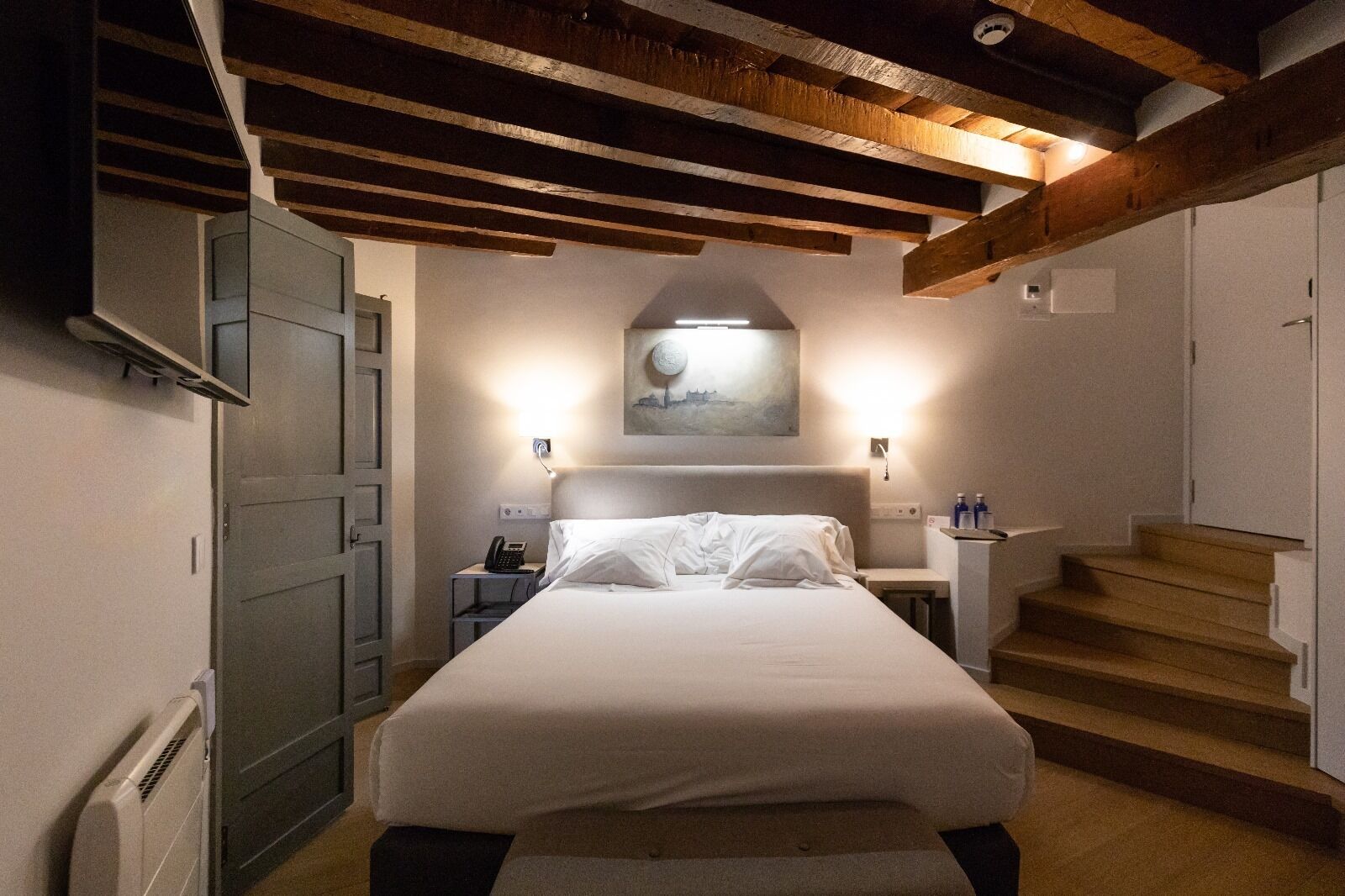 A bedroom with a bed, bedside tables, a phone, a painting, and a staircase.