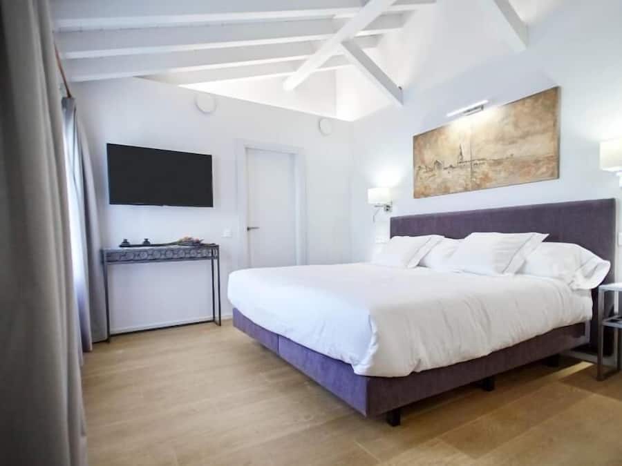 Una habitación de hotel moderna con una cama grande, un televisor de pantalla plana montado en la pared, un cuadro sobre la cama y un escritorio con una lámpara.