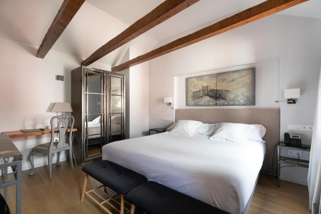 Un dormitorio con cama, escritorio, silla, banco, armario y un cuadro en la pared.