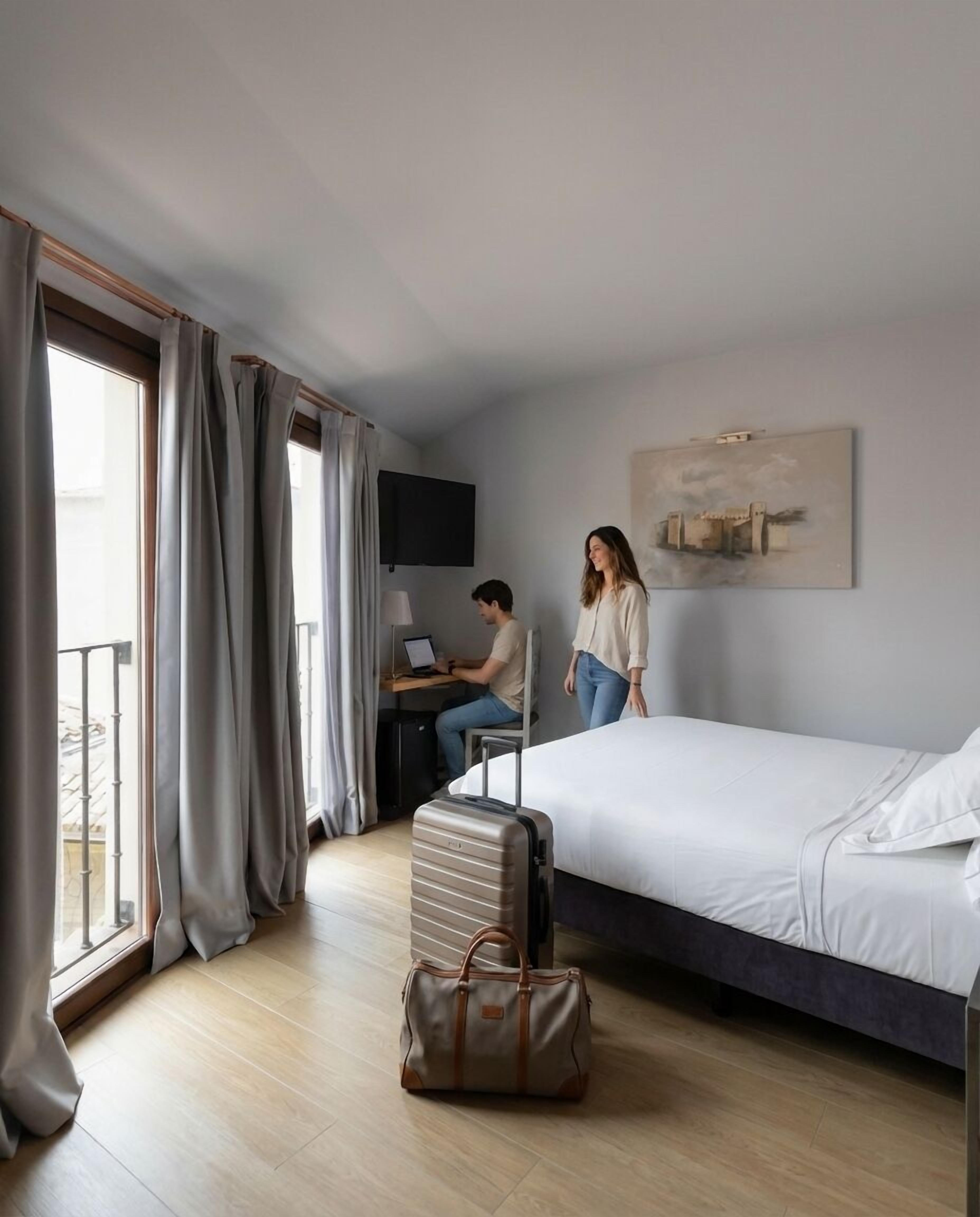 A hotel room with a bed, a desk with a laptop, a suitcase, and a painting on the wall.