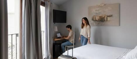 A hotel room with a bed, a desk with a laptop, a suitcase, and a painting on the wall.