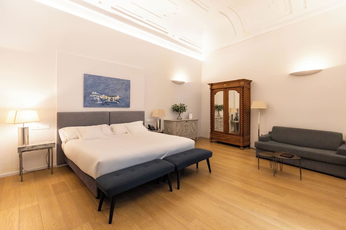 Une chambre d’hôtel moderne avec un grand lit, un canapé gris, une petite table, un miroir et un tableau au mur.