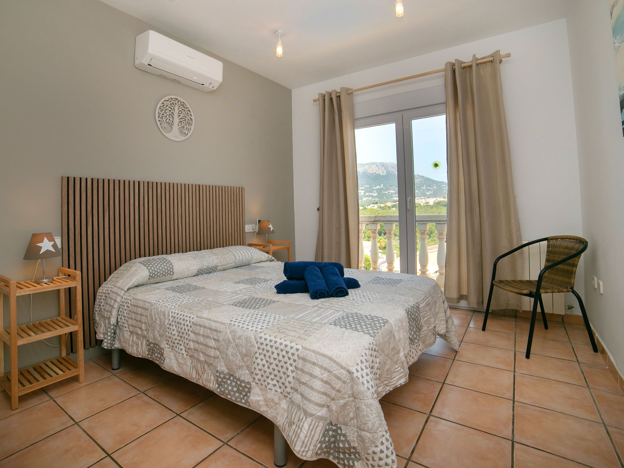 A bedroom with a bed, a chair, a nightstand, a lamp, a window with curtains, and an air conditioning unit.