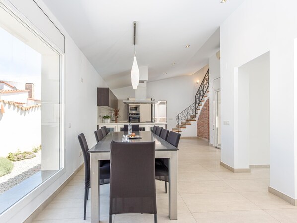 Een moderne eetruimte met een grote tafel en stoelen, een keuken op de achtergrond, en een trap naar een hoger niveau.