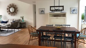 Un salon moderne avec un canapé, une table basse et des fauteuils. Une table à manger avec des chaises se trouve au premier plan. Un miroir décoratif est accroché au mur et une œuvre d'art est placée au-dessus de la table à manger.