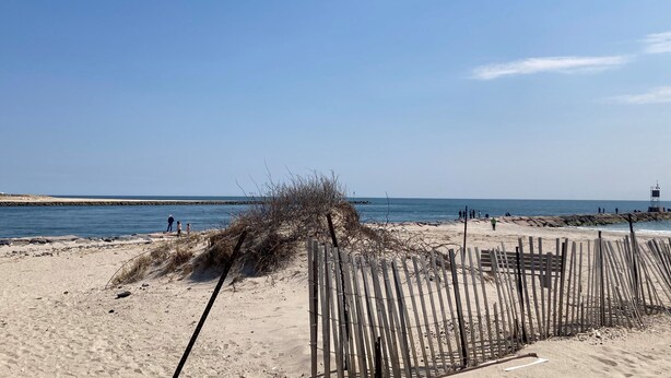 Une plage de sable bordée d’une clôture en bois, avec une digue rocheuse en arrière-plan.