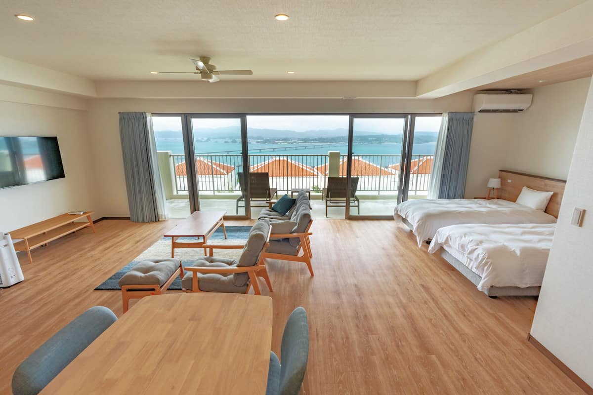 A hotel room with a large balcony, a bed, a sofa, a TV, and a dining table.