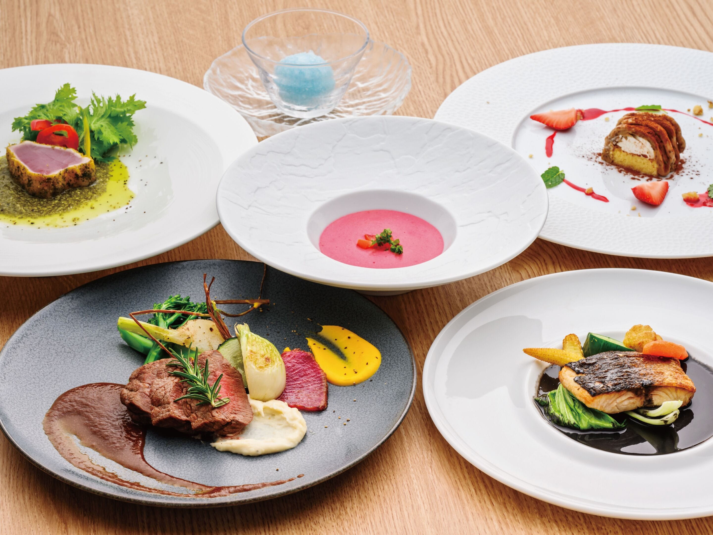 Un tavolo imbandito con diversi piatti, tra cui una zuppa rosa, una bistecca e un piatto di pesce.