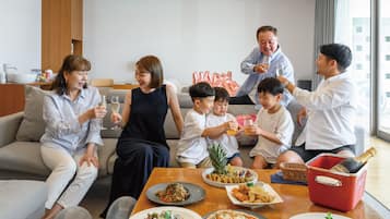 A group of people seated around a table with various dishes and drinks, in a modern living room with a sofa and a wooden cabinet.