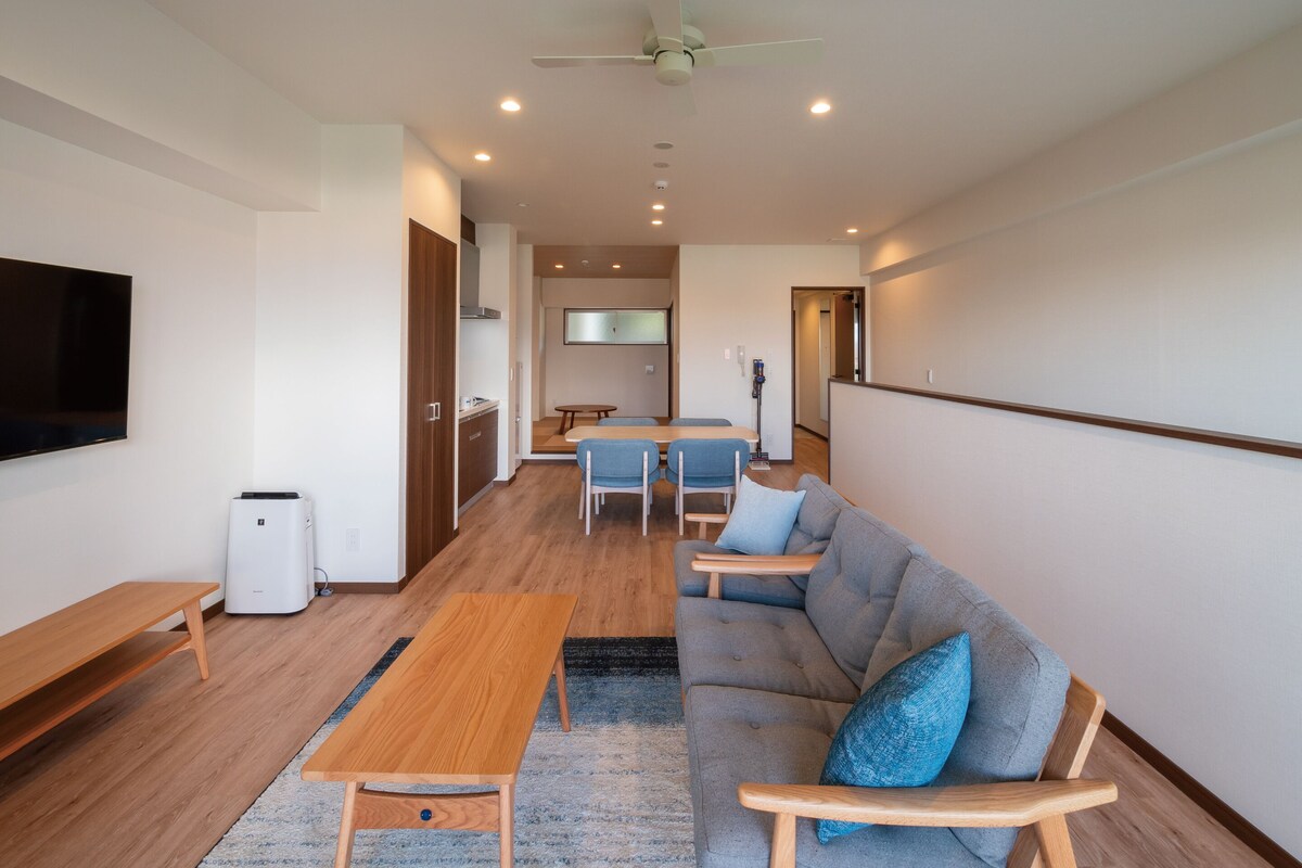 A modern living room with a sofa, a coffee table, a TV, and a kitchen area in the background.