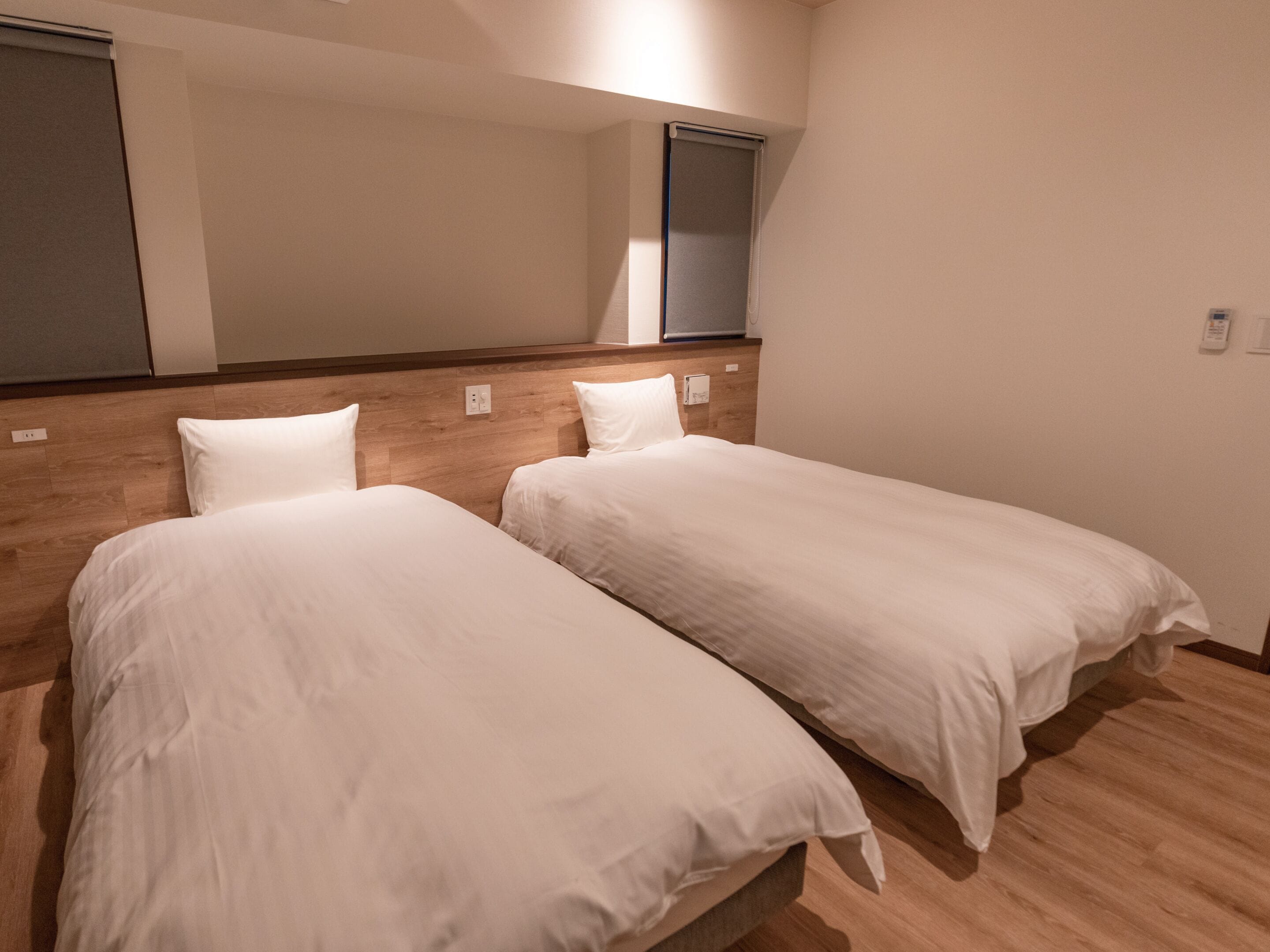 A hotel room with two single beds, wooden headboards, a large mirror, and a window with blinds.