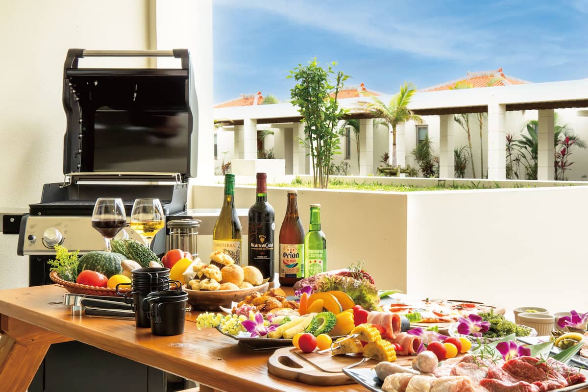 A barbecue setup with a grill, various food items, and drinks on a table, overlooking a resort with palm trees and buildings.