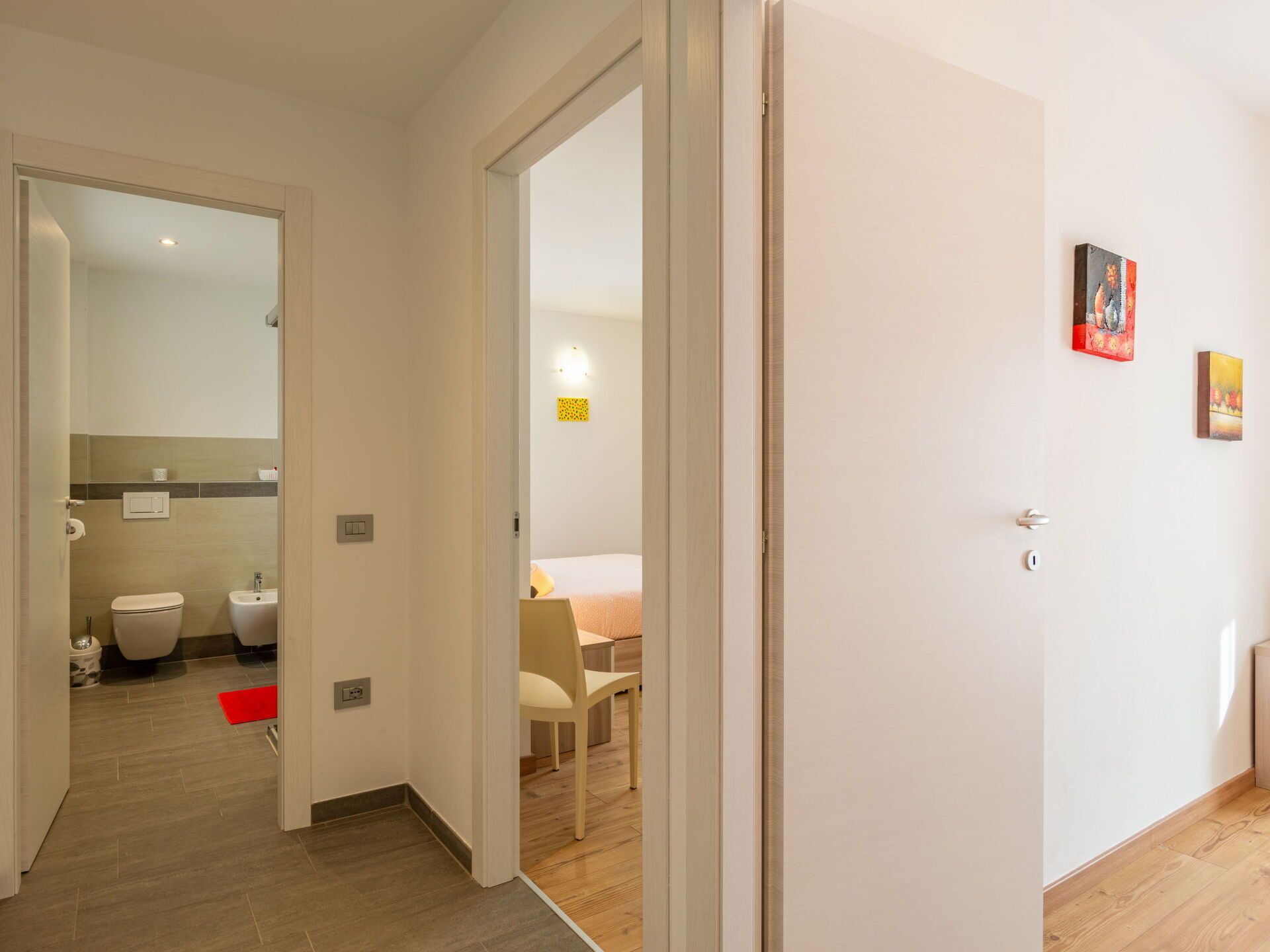 Une chambre d’hôtel avec un plancher en bois, un lit, une chaise et une petite table. Elle comprend une salle de bain avec des toilettes et un lavabo.