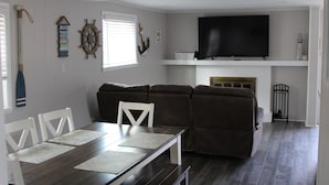 A living room with a dark wooden dining table, white chairs, a brown sectional sofa, a flat-screen TV mounted above a fireplace, and nautical decor on the walls. - Updated & short walk to downtown Charlevoix! (Charlevoix)