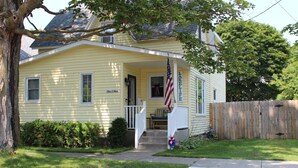 A yellow house with a porch, an American flag, and a wooden fence. - Updated & short walk to downtown Charlevoix! (Charlevoix)