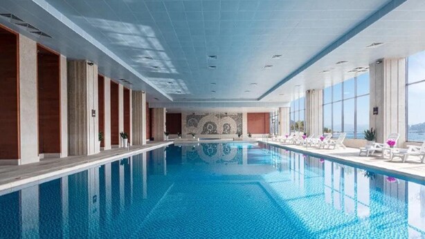 Una amplia piscina interior con agua cristalina, rodeada de sillones de descanso y vistas al mar.